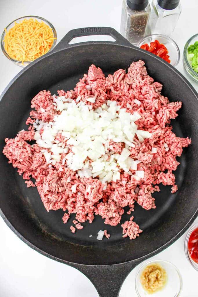 A skillet filled with raw ground beef and chopped onions, surrounded by bowls containing shredded cheese, diced tomatoes, chopped green vegetables, minced garlic, and seasonings&mdash;perfect for whipping up a delicious cheeseburger pasta.