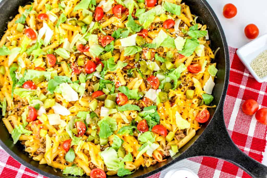 A skillet containing a cheeseburger pasta dish topped with shredded cheese, chopped lettuce, cherry tomatoes, and diced pickles. The skillet is placed on a red and white checkered cloth, with extra ingredients nearby.