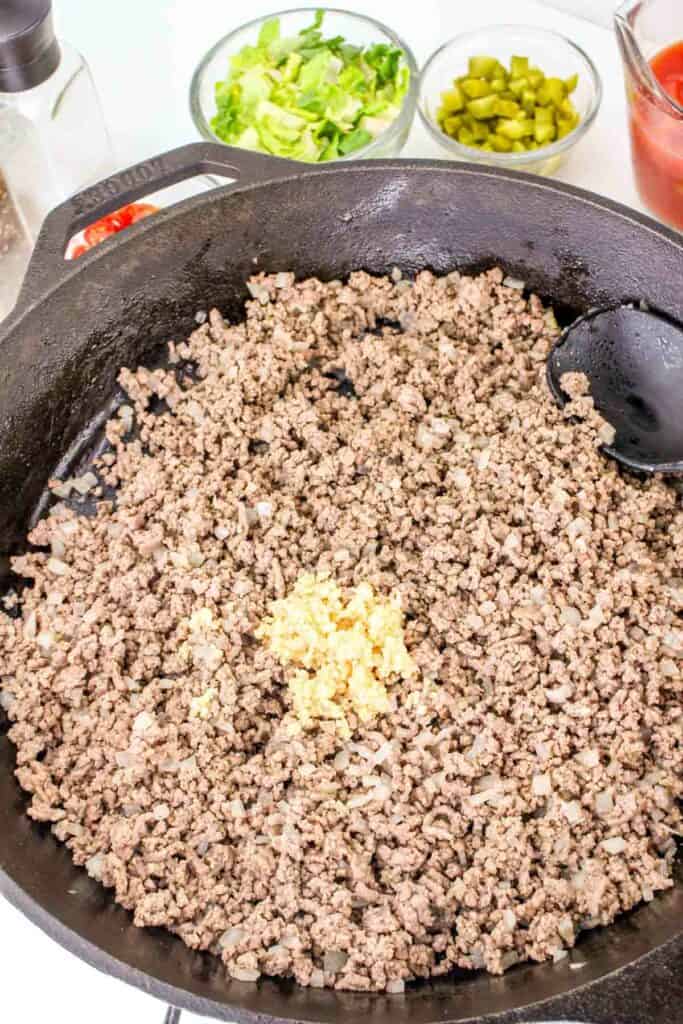 A cast-iron skillet contains browned ground beef and minced garlic, just like the base for a delicious cheeseburger pasta. Bowls with chopped lettuce, pickles, and tomatoes are in the background. A black spoon rests in the skillet, ready for serving.