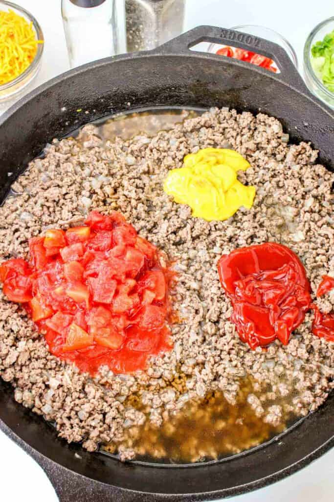 A black skillet with cooked ground beef topped with diced tomatoes, ketchup, and mustard. In the background are bowls of shredded cheese, sliced green onions, and seasonings, perfect for creating a delicious cheeseburger pasta.