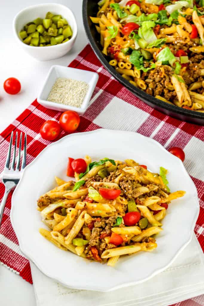 A plate of cheeseburger pasta mixed with ground beef, chopped vegetables, and garnished with cherry tomatoes sits on a red and white tablecloth. A bowl of pickles and a dish of sesame seeds are beside the plate.