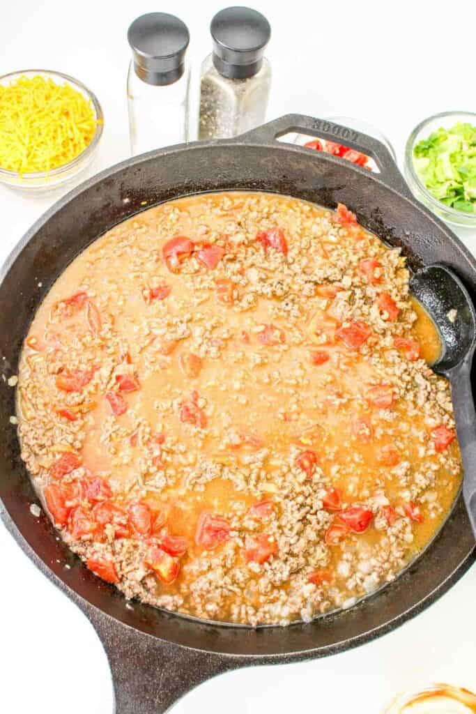 A cast iron skillet filled with a ground meat and tomato mixture reminiscent of cheeseburger pasta, surrounded by small bowls containing grated cheese, chopped greens, salt, and pepper.