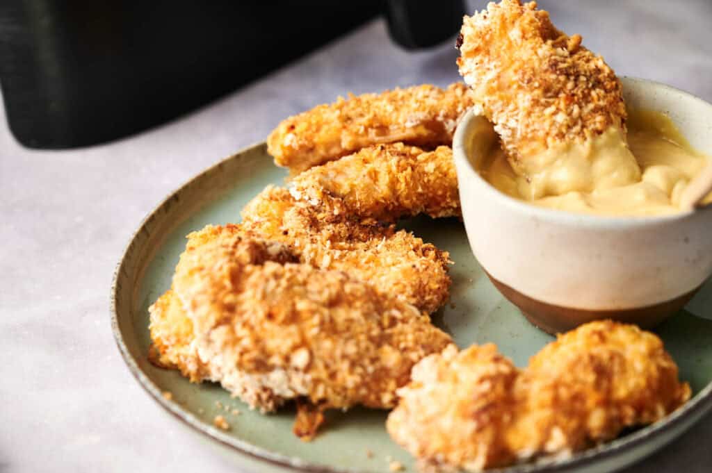 A plate of air fryer chicken tenders with one piece dipped in a bowl of yellow sauce.