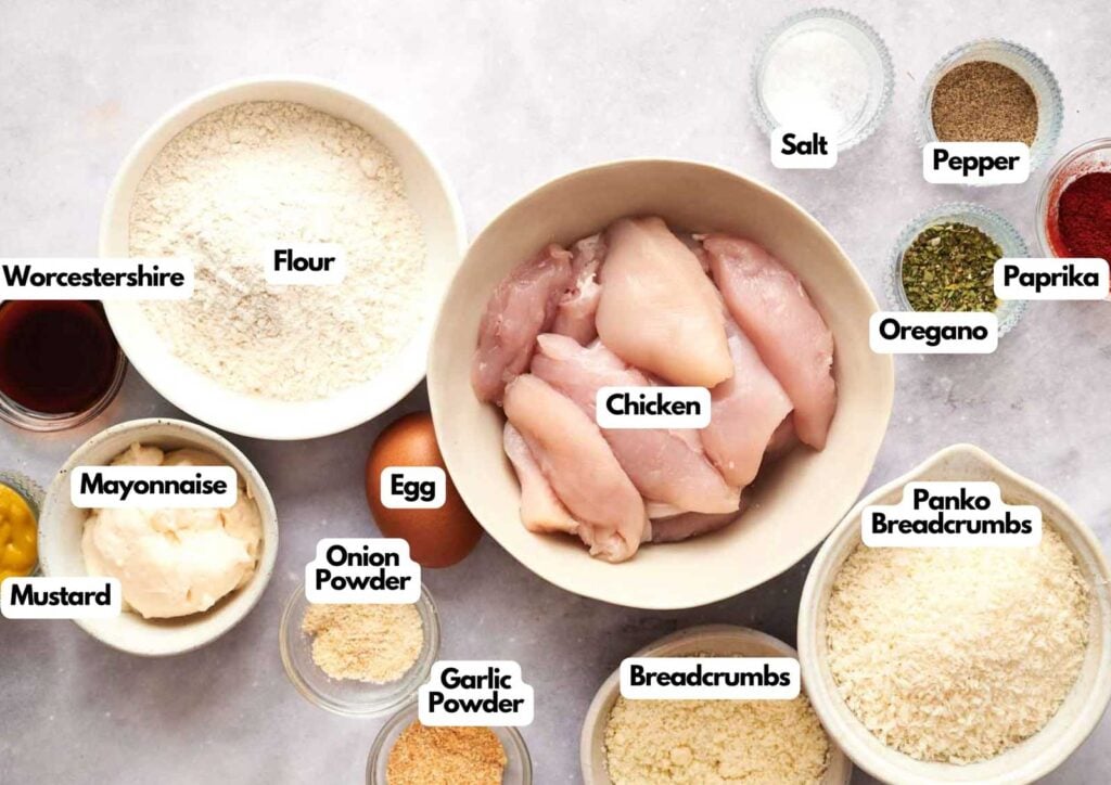 Overhead view of labeled ingredients in bowls, ready for Air Fryer Chicken Tenders&mdash;raw chicken, flour, panko breadcrumbs, egg, mayonnaise, mustard, Worcestershire sauce, spices, salt, and pepper.
