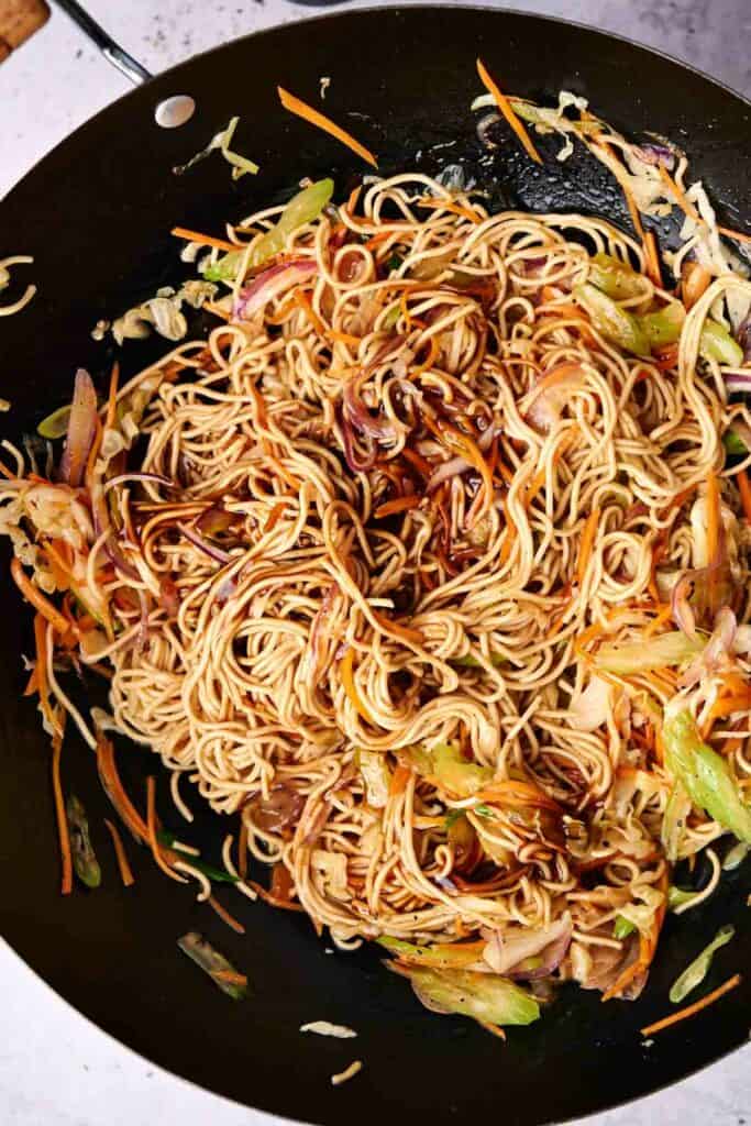 A wok containing stir-fried chow mein noodles mixed with vegetables like shredded carrots, cabbage, and onions.