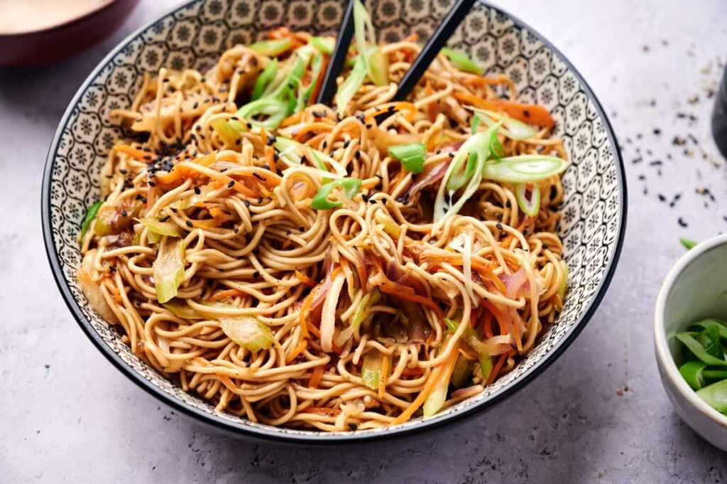 A bowl of chow mein stir-fried noodles with sliced vegetables, garnished with green onions and black sesame seeds, with a pair of chopsticks resting in the bowl.