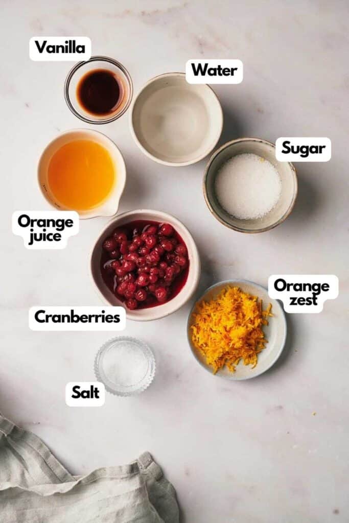 Ingredients for a recipe are laid out on a marble surface labeled as vanilla, water, sugar, orange juice, cranberries, orange zest, and salt.