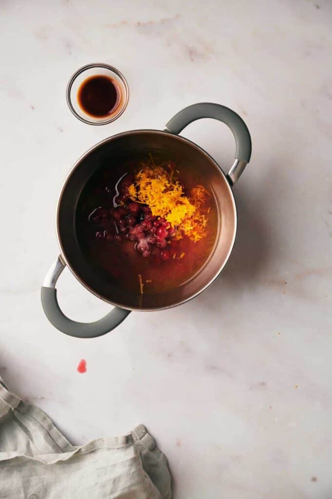 A saucepan containing ingredients including cranberries and orange zest on a marble surface with a small glass bowl of liquid beside it. A cloth is partly visible at the bottom left corner.