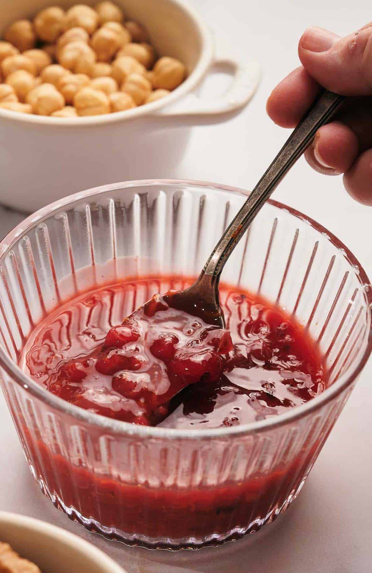 A hand holding a spoon with raspberry jam in a glass bowl, with hazelnuts in a bowl in the background.