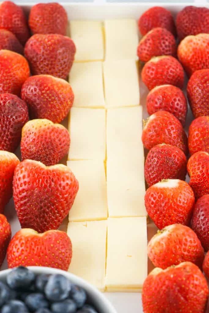 A tray containing rows of strawberries and squares of cheese in the center, with a bowl of blueberries in the foreground.