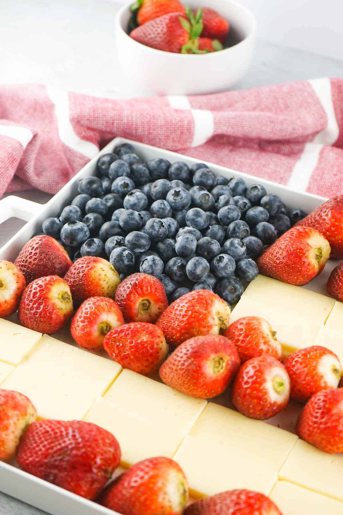 A rectangular tray with strawberries, blueberries, and rectangular slices of cheese arranged to resemble the American flag. A white bowl of strawberries and a red-striped cloth are in the background.