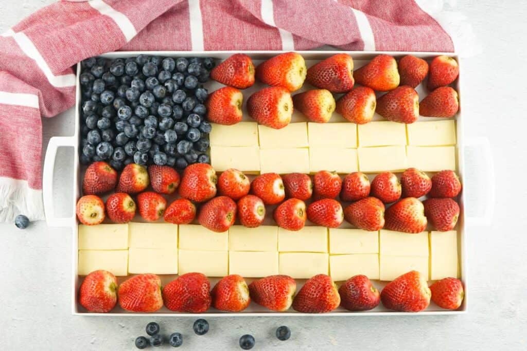 A rectangular tray of dessert arranged to resemble the American flag, with blueberries in the top left corner and alternating rows of strawberries and cream squares.