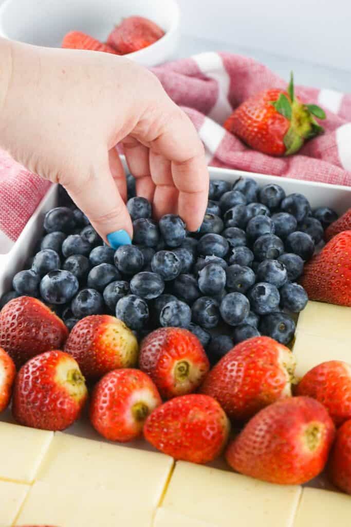 A hand with blue-painted nails reaches for blueberries from a tray. The tray also contains strawberries and cheese cubes.