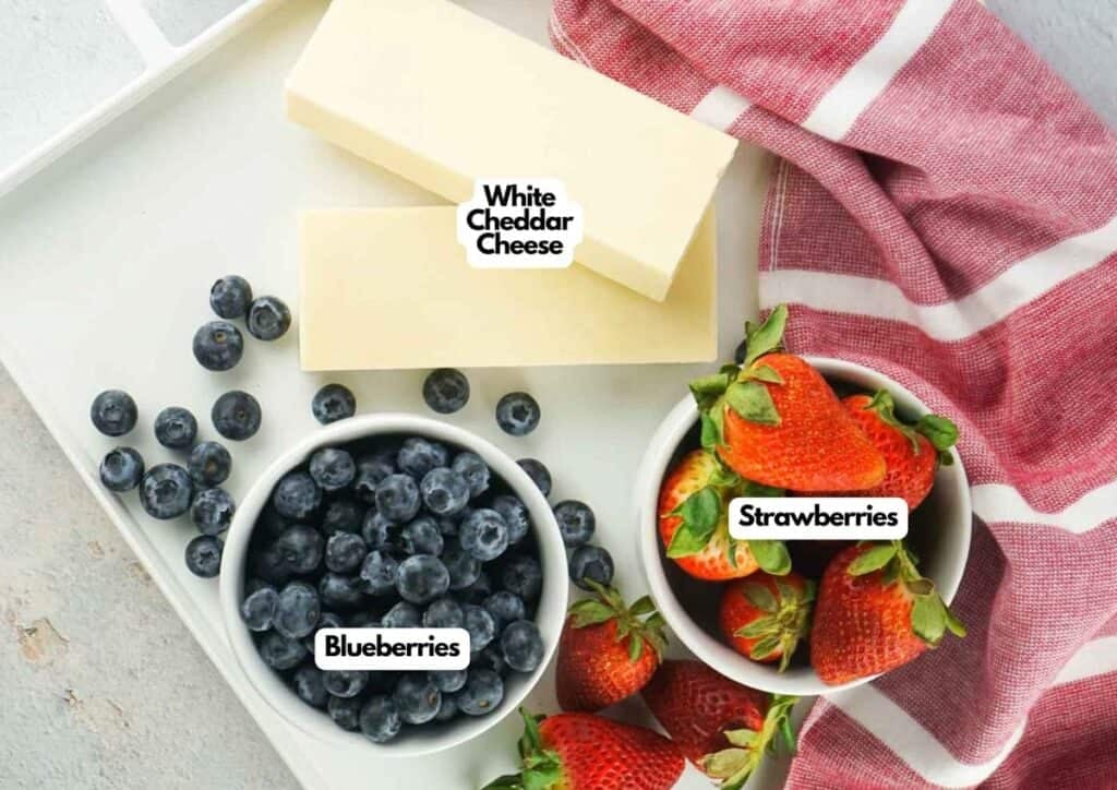 A bowl of blueberries, a bowl of strawberries, and two blocks of white cheddar cheese on a tray with a red and white towel.