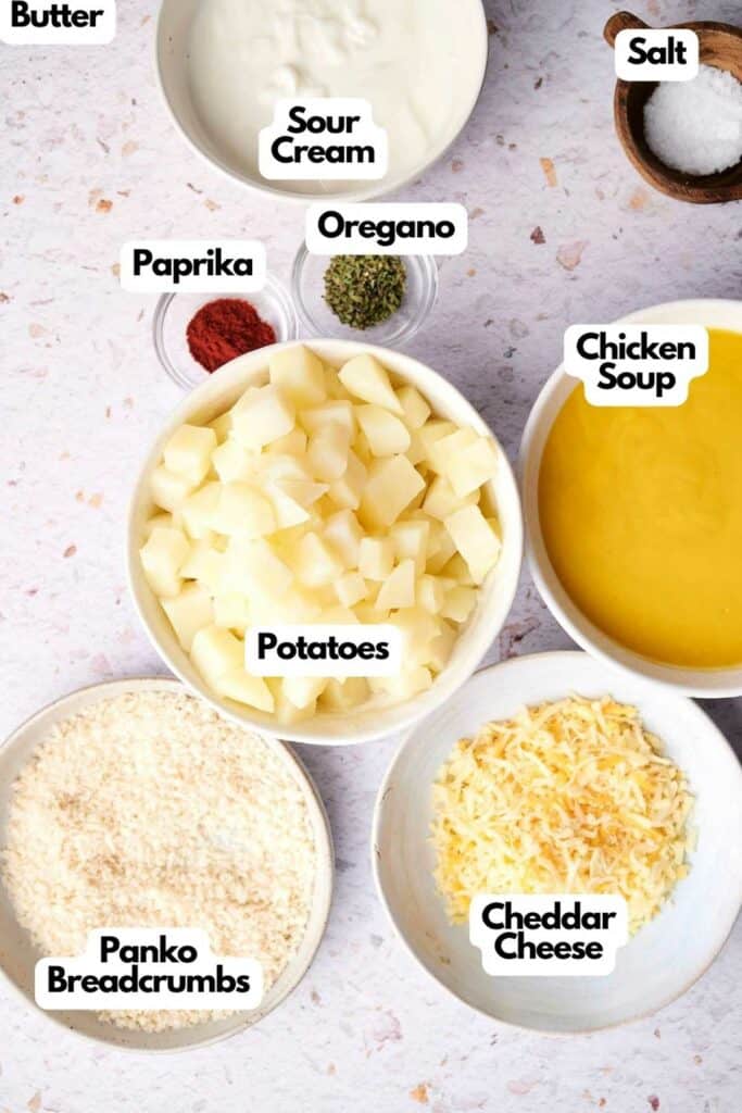 Ingredients for a recipe, including cubed potatoes, shredded cheddar cheese, chicken soup, panko breadcrumbs, butter, sour cream, paprika, oregano, and salt&mdash;everything you need to make classic funeral potatoes&mdash;arranged in bowls.
