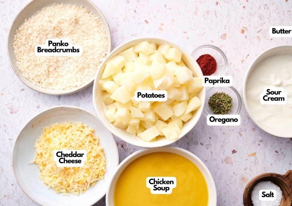 Bowls containing the classic funeral potatoes ingredients—panko breadcrumbs, diced potatoes, cheddar cheese, chicken soup, paprika, oregano, butter, sour cream, and salt—arranged on a surface.