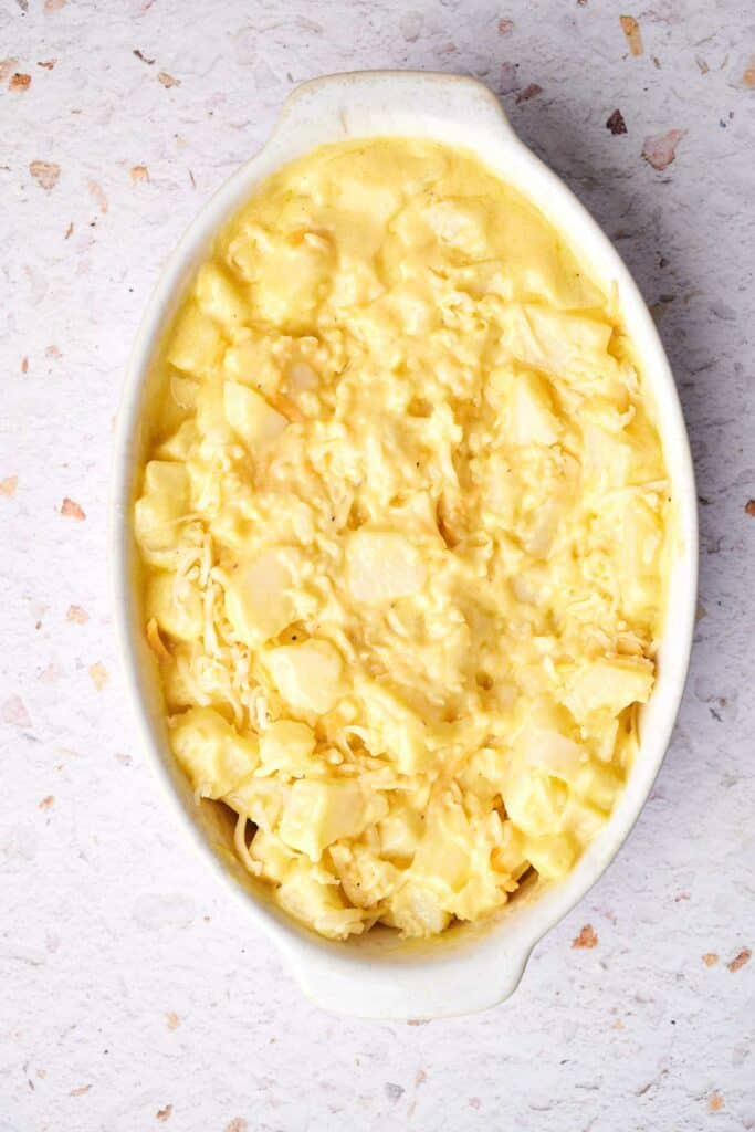 A white oval dish filled with cheesy, creamy funeral potatoes on a light textured surface.
