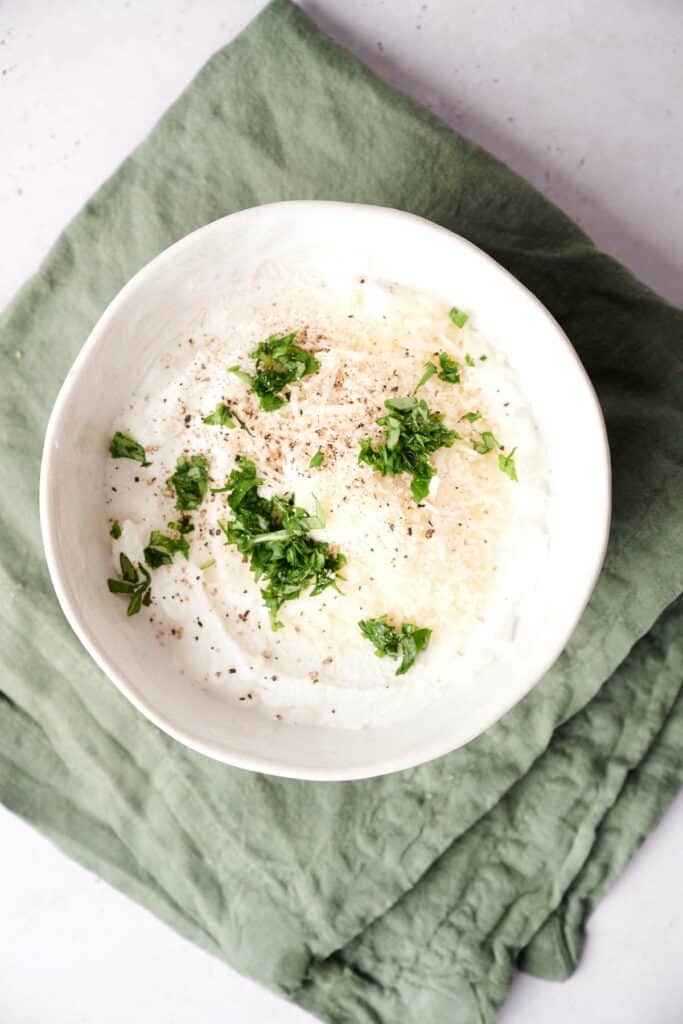 A white bowl filled with a creamy lasagna mixture, topped with grated cheese, black pepper, and chopped parsley, placed on a green cloth.