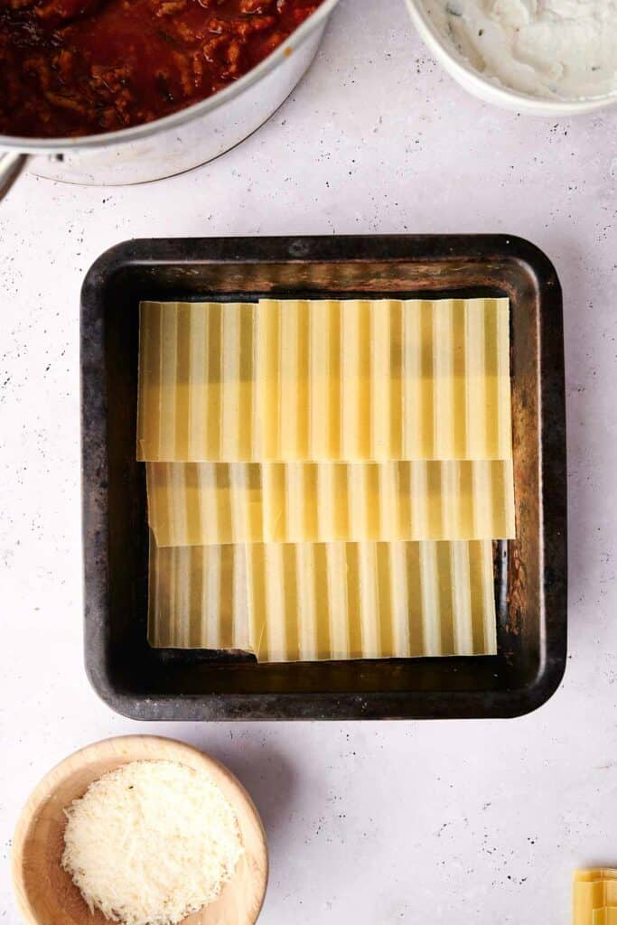 A square baking pan filled with layered uncooked lasagna sheets, ready to become a delicious lasagna. Nearby, a bowl of grated cheese and a pot of rich tomato-based sauce wait to complete the meal.