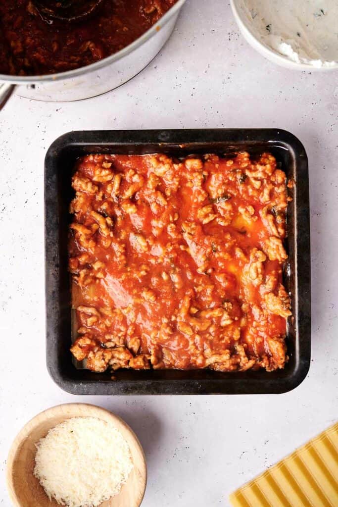 A black baking dish filled with a delicious layer of lasagna ingredients, primarily consisting of tomato sauce and ground meat, sits on a countertop next to a bowl of grated cheese and a pot of red sauce.