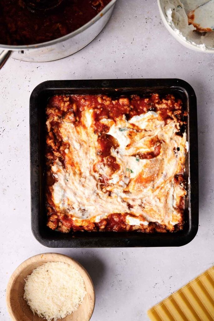 A square baking pan filled with unbaked lasagna, layered generously with tomato sauce and cheese on top. Nearby are a bowl of grated cheese, uncooked lasagna noodles, and a pot of rich lasagna sauce.