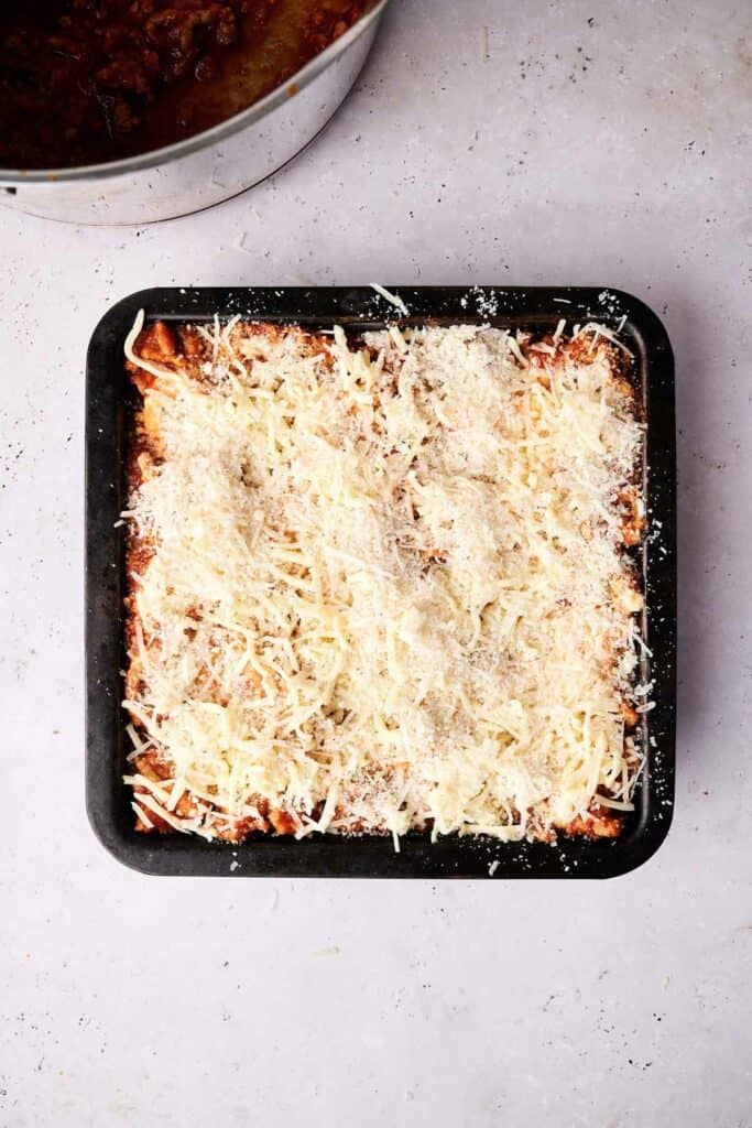 A square baking dish filled with an uncooked lasagna topped with shredded cheese. A portion of a pan with sauce is visible in the top left corner. The surface of the lasagna is whitish grey.