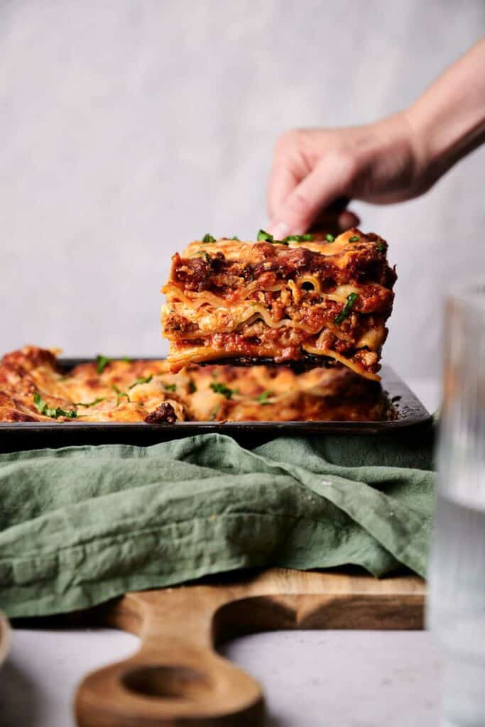A hand lifting a square piece of lasagna from a pan, revealing delicious layers of pasta, cheese, and sauce. The pan rests on a green cloth and wooden board. A glass of water is visible in the foreground, completing the inviting scene.
