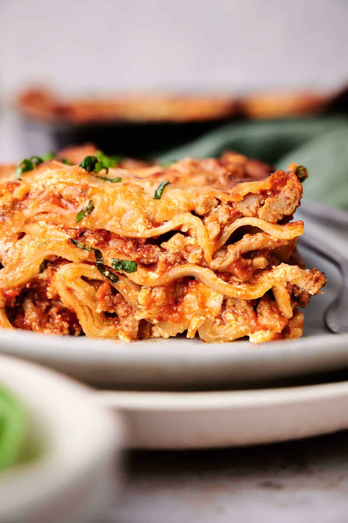Close-up of a mouthwatering slice of lasagna on a gray plate, showcasing its layers of tender pasta, savory meat, creamy cheese, and rich sauce, all garnished with fresh chopped herbs.
