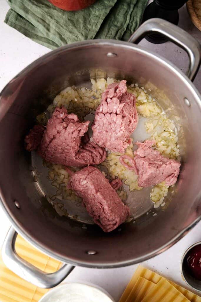 Ground meat and chopped onions sizzle in a large stainless steel pot, perfectly prepped for a delicious lasagna, on a white kitchen counter.