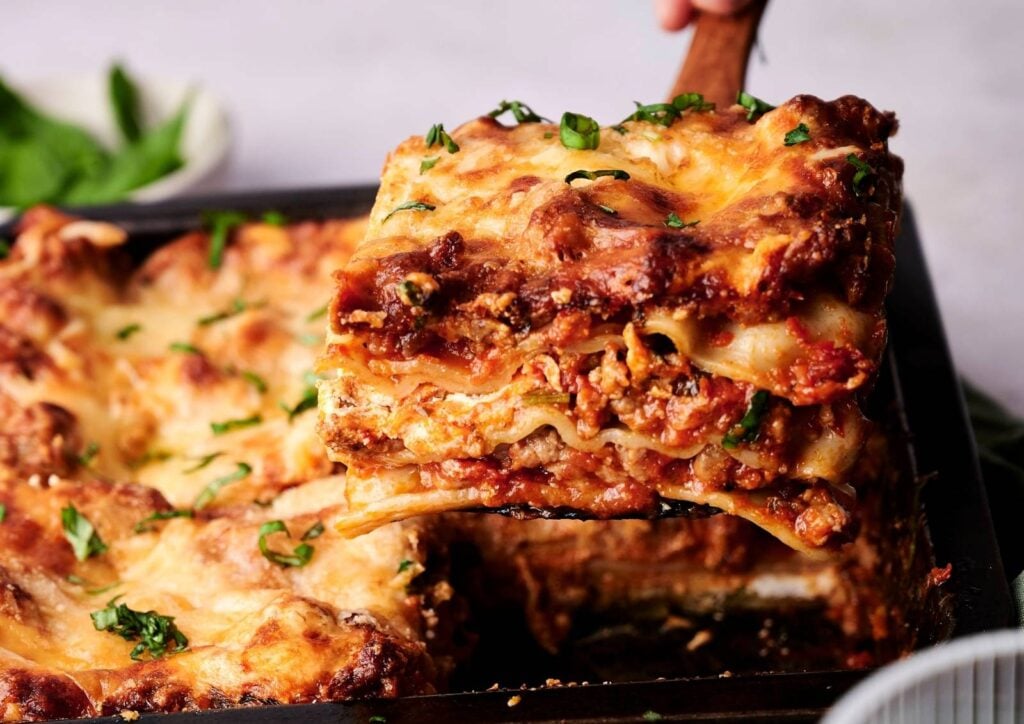 A slice of cheesy, baked lasagna with layers of tender pasta, rich meat sauce, and gooey melted cheese being lifted from a pan. This classic lasagna is the ultimate comfort food, perfect for sharing at family dinners.