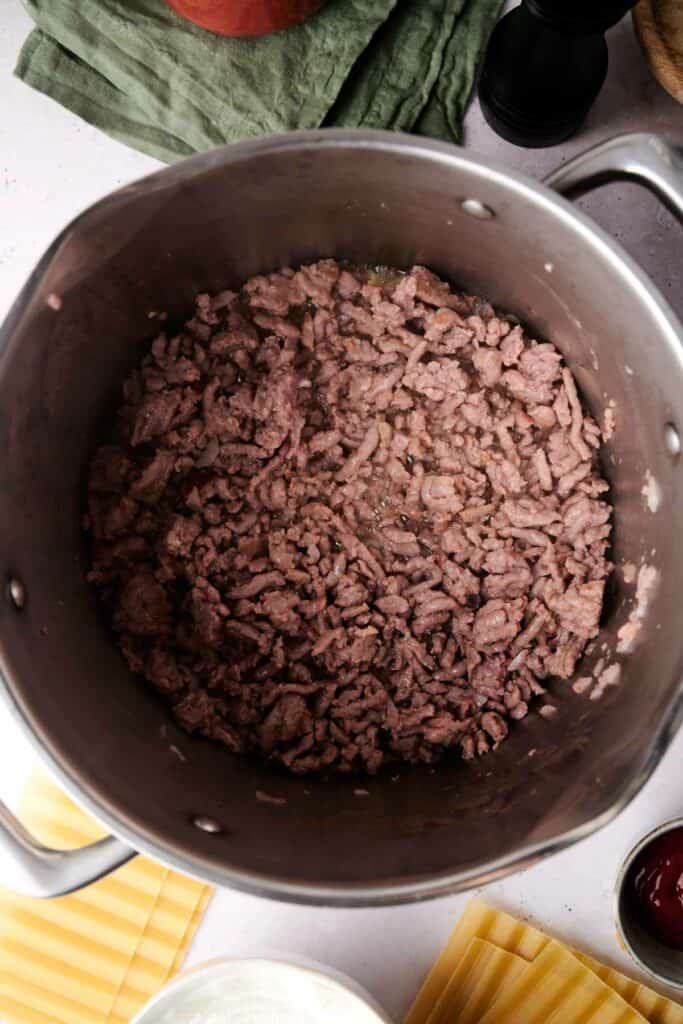 A pot filled with cooked, browned ground meat sits on a countertop with a green cloth, pasta sheets, and other kitchen items nearby, ready to assemble a delicious lasagna.