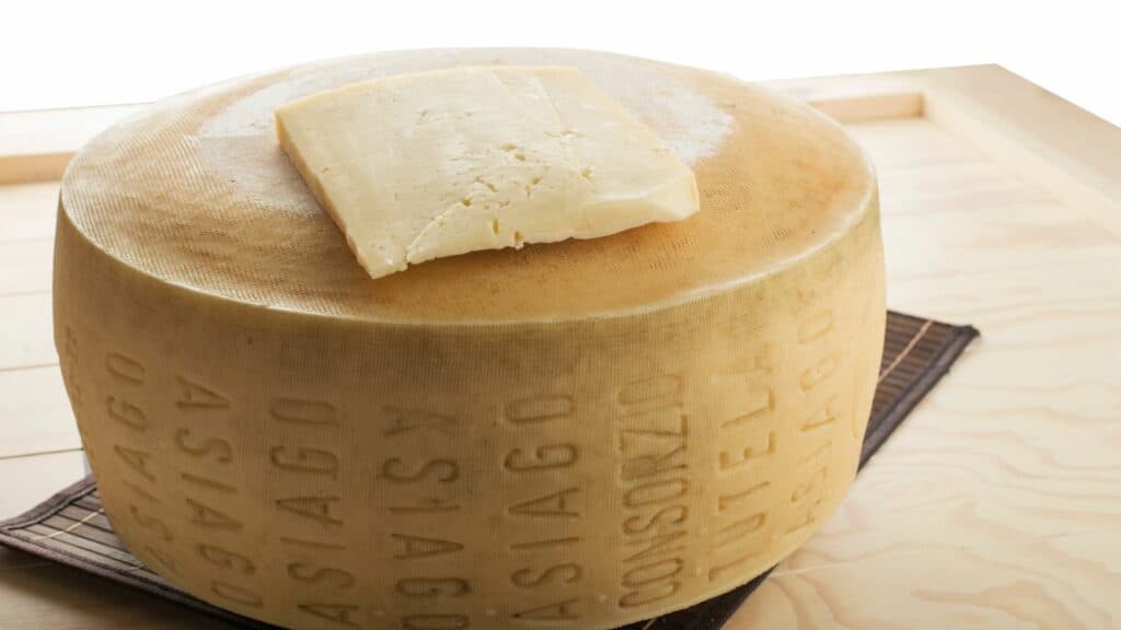 A large wheel of Asiago cheese with a smaller rectangular slice on top, displayed on a wooden surface.