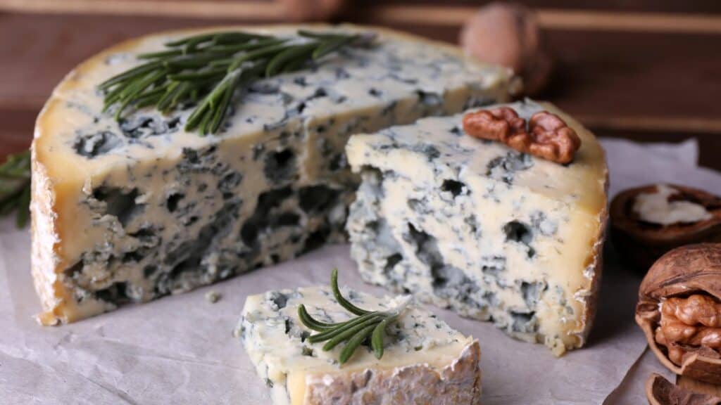 A wedge of blue cheese garnished with sprigs of rosemary and surrounded by walnuts on a wooden board.