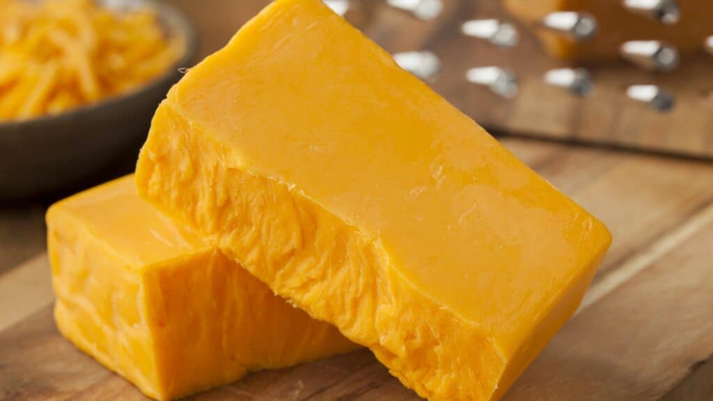 Two blocks of cheddar cheese on a wooden surface. A cheese grater is visible in the background.