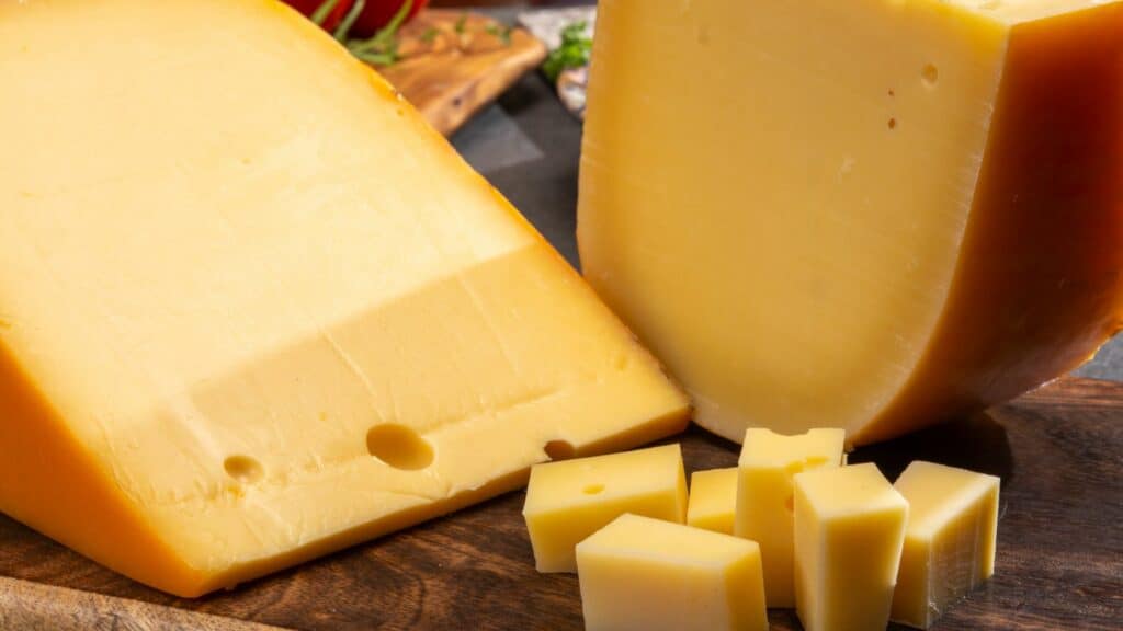 A close-up of a block of yellow cheese partially sliced, with several small cubes of the same cheese in the foreground, placed on a wooden surface.