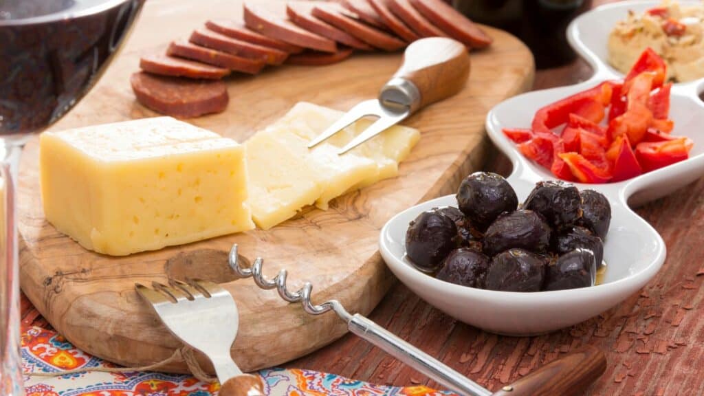 A wooden platter with sliced cheese and sausage, accompanied by bowls of black olives and red bell peppers. A cheese fork and corkscrew are also included on the table.