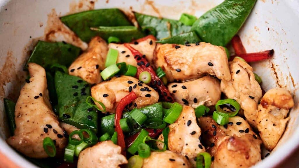 A close-up of a chicken stir-fry dish with snow peas, red bell peppers, green onions, and black sesame seeds.