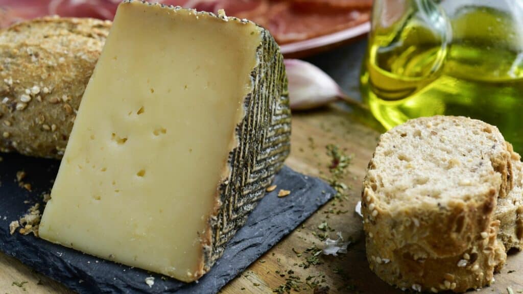 A piece of hard cheese and slices of bread are displayed on a slate board, with olive oil and a garlic clove in the background.