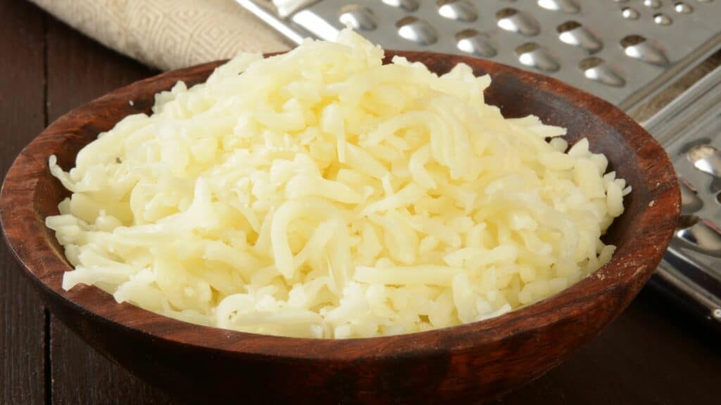 A wooden bowl filled with shredded cheese sits on a wooden surface next to a metal grater.