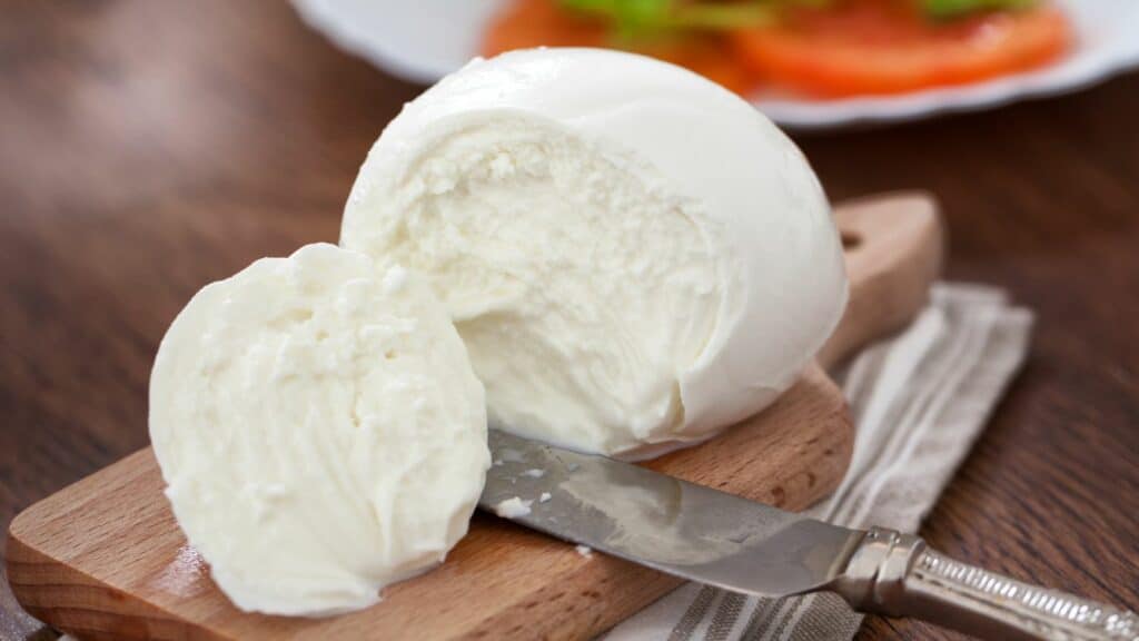A ball of fresh mozzarella cheese on a wooden cutting board with a knife and cloth napkin. A portion of the cheese is sliced off. A plate with sliced tomatoes is blurred in the background.