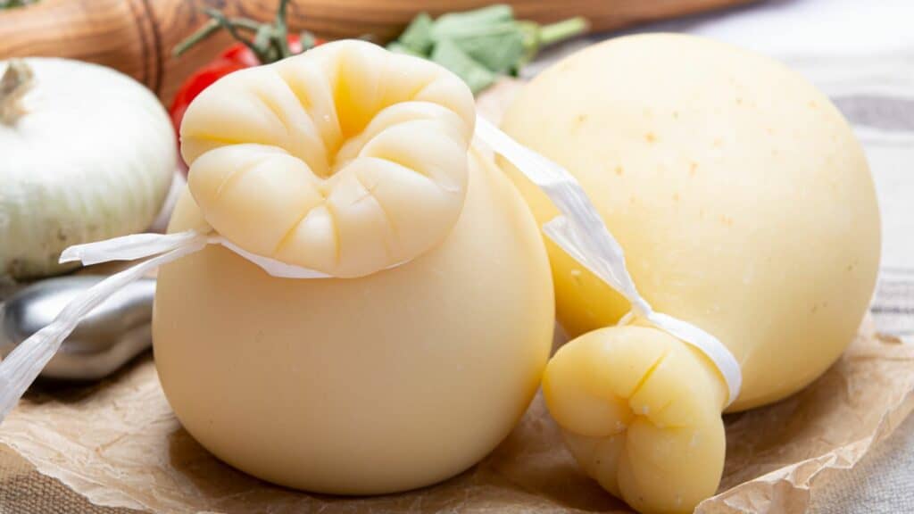 Two round cheese wheels with knotted tops and ribbons, placed on brown paper, surrounded by garlic, herbs, and a tomato in the background.