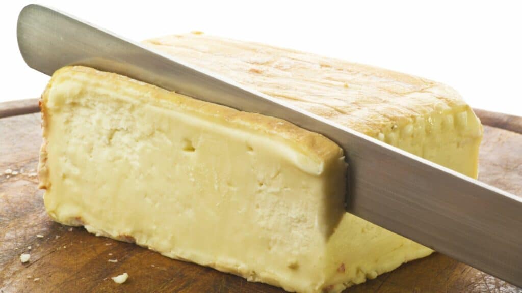 A knife slicing through a block of cheese on a wooden cutting board.