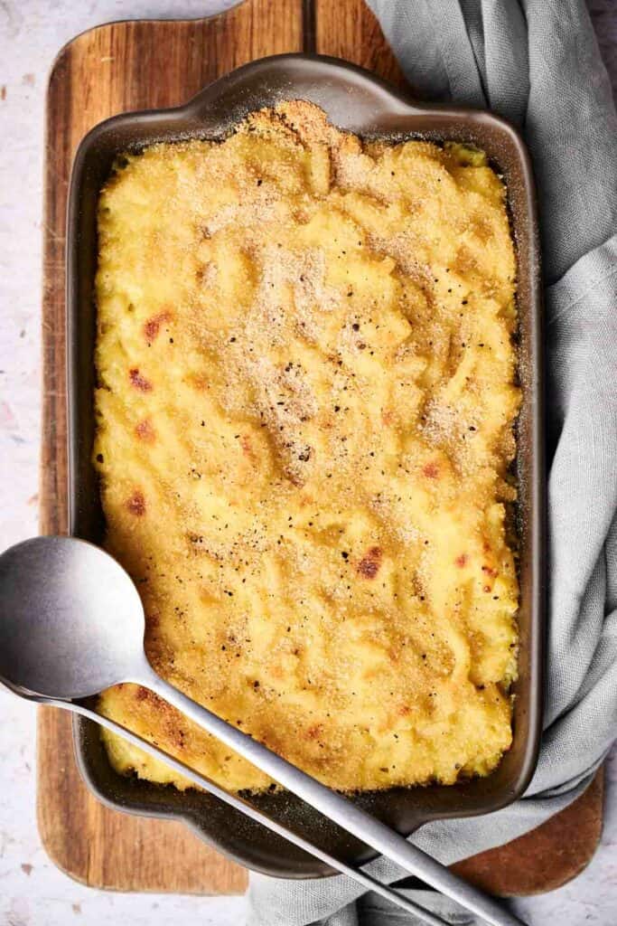 A rectangular baking dish filled with baked mac and cheese, sprinkled with breadcrumbs next to a gray cloth and a metal spoon.