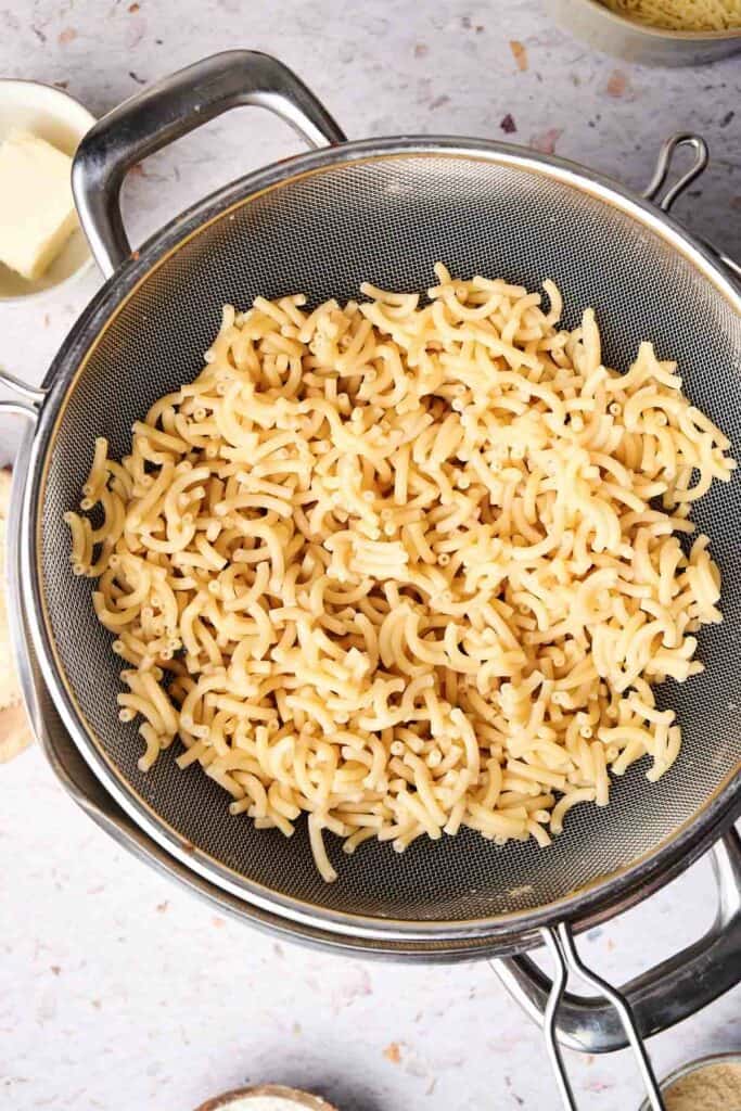 Cooked macaroni pasta in a metal colander, draining over a pot, with cups of butter and grated cheese visible in the background, ready to transform into creamy mac and cheese.