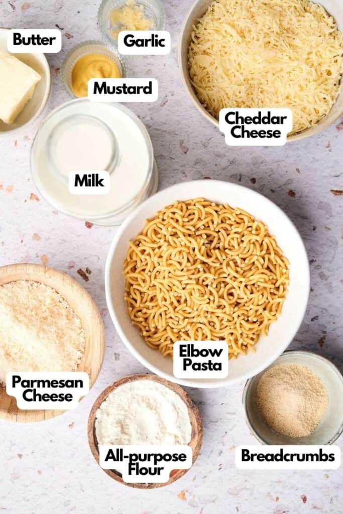 Image of various ingredients labeled on a countertop: elbow pasta, milk, cheddar cheese, garlic, mustard, butter, parmesan cheese, all-purpose flour, and breadcrumbs—everything you need for a perfect mac and cheese.