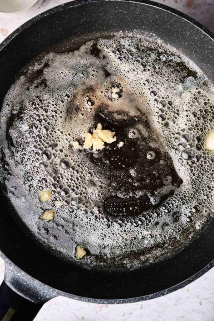 A black skillet with foaming butter, small chunks of garlic browning in it, sets the perfect base for a creamy mac and cheese.