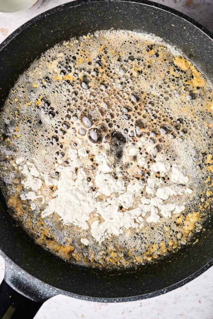 A frying pan with bubbling brown sauce and scattered white flour on top, cooking on a stovetop, hints at the delicious promise of homemade mac and cheese.