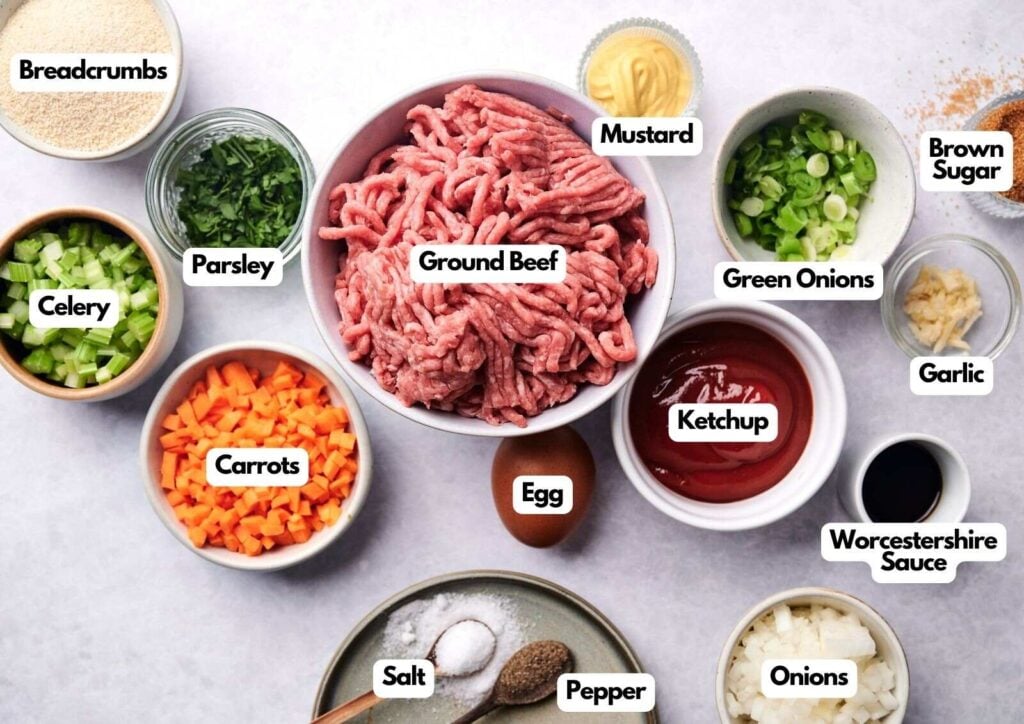 A variety of labeled meatloaf ingredients are arranged on a white surface, including ground beef, ketchup, egg, vegetables, breadcrumbs, and seasonings in bowls.