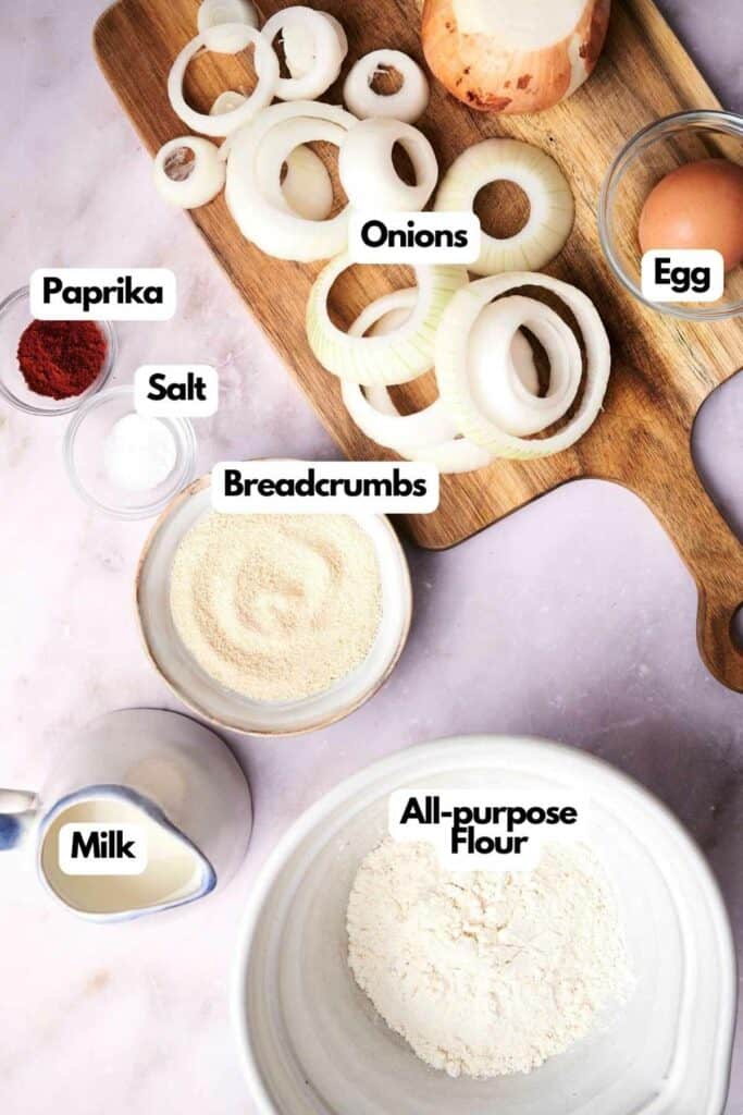 Ingredients for mouth-watering onion rings include fresh onion slices on a wooden board, an egg, paprika, salt, breadcrumbs, all-purpose flour, and milk.