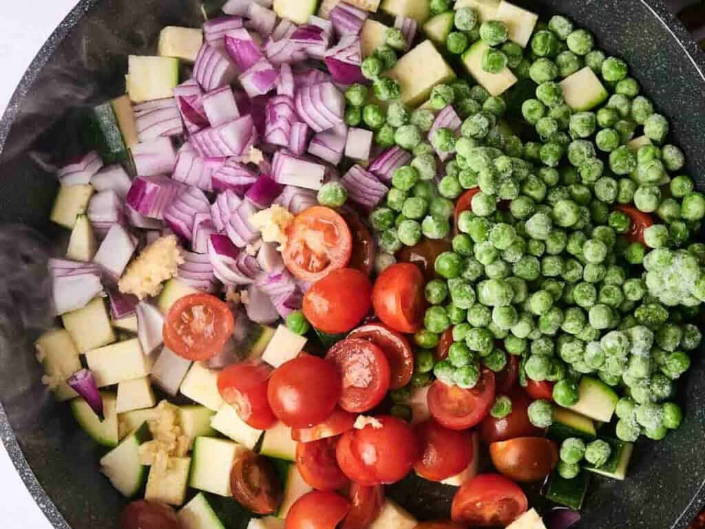 A pan filled with chopped red onions, peas, cherry tomatoes, zucchini, and minced garlic. A jar of oil and some asparagus are nearby.